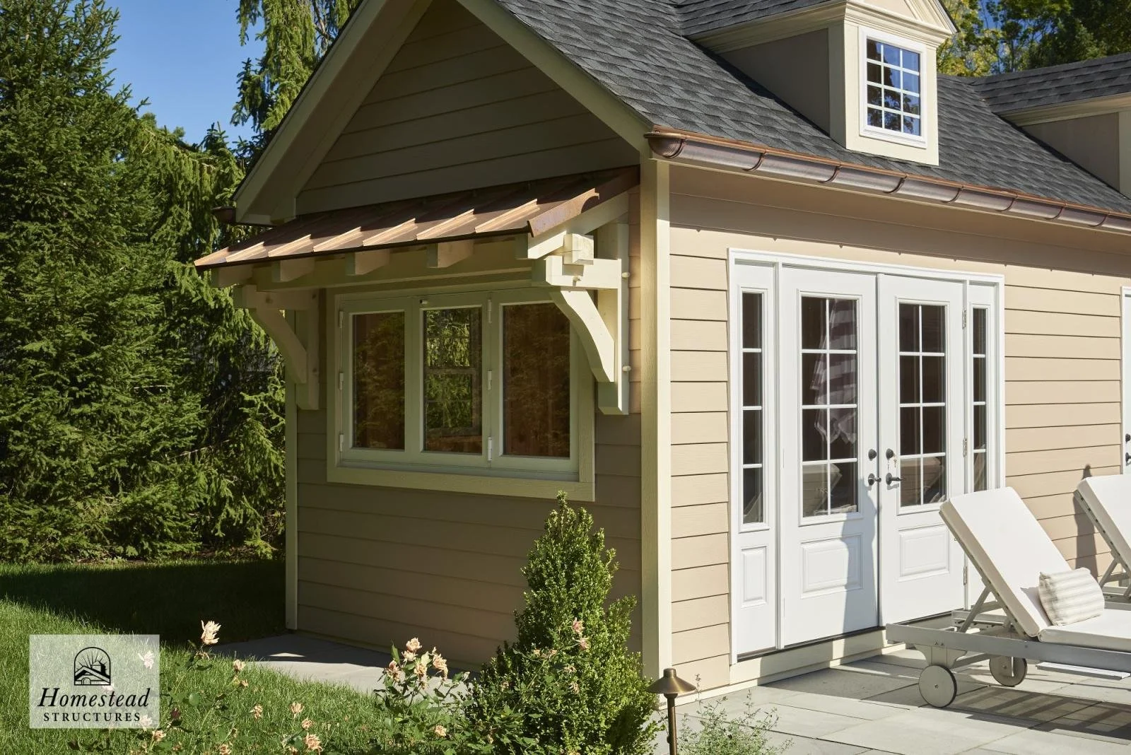 Exterior view of a beige house with white double doors, a window with shutters, and a small gabled roof with dormer window, surrounded by greenery and lawn chairs.