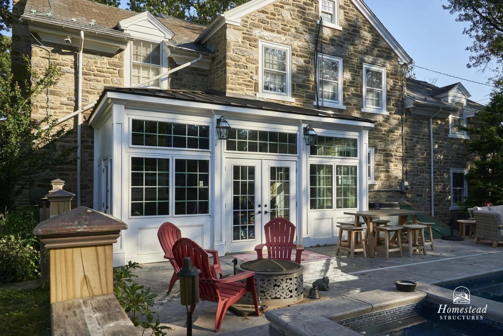 A backyard patio area with red Adirondack chairs, a fire pit, and a table with stools. The patio is in front of a stone house with a white sunroom extension featuring large windows and double doors. The house has multiple windows and a pitched roof.