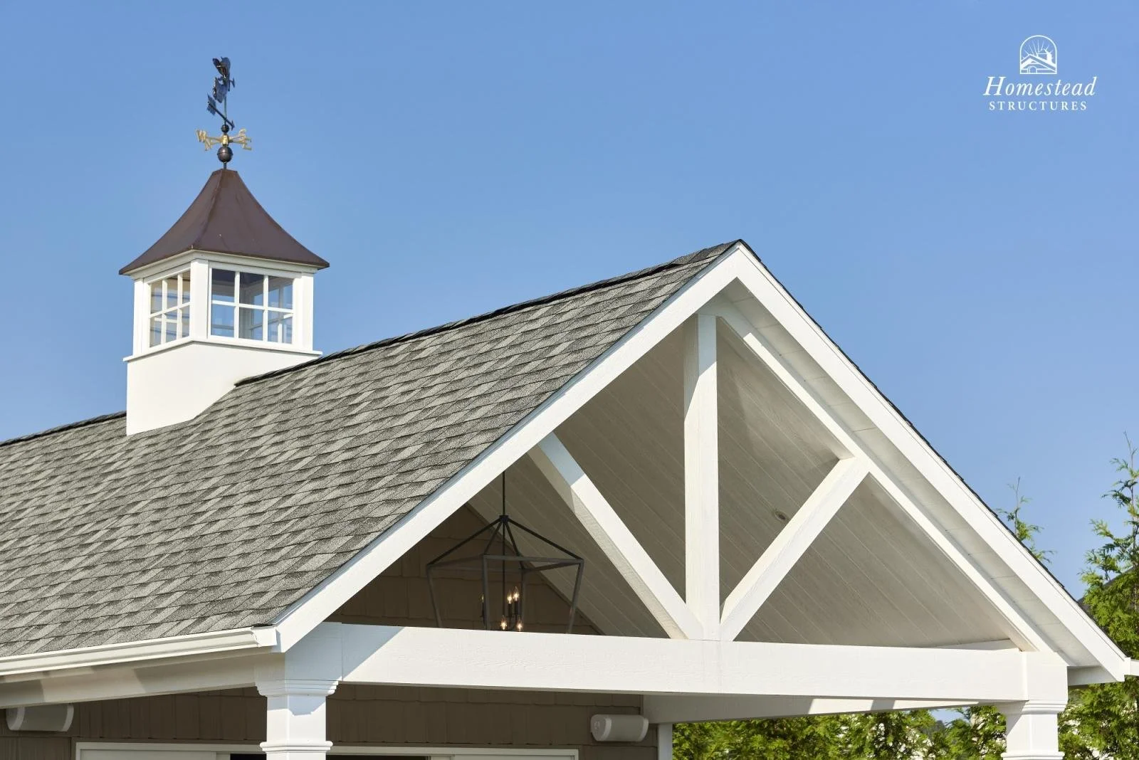 Close-up of a house roof with a cupola and weather vane against a clear blue sky, with a logo for Homestead Structures in the top right corner.