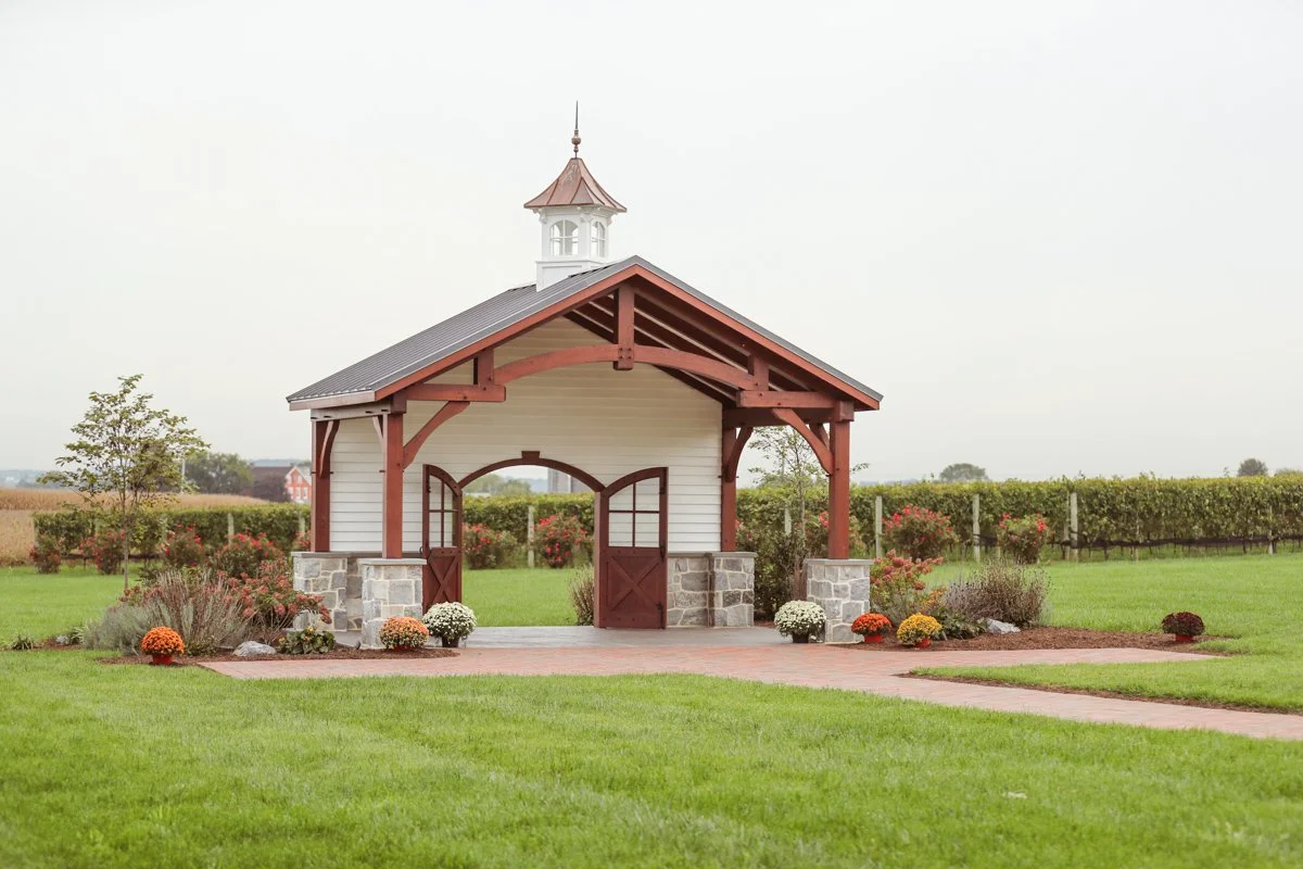 A small, white and brown pavilion with a stone foundation, located on a well-manicured lawn with flowers and shrubs, overlooking fields.