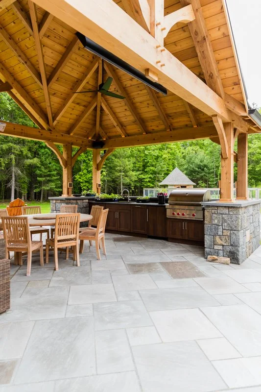 Outdoor kitchen and dining area with a wooden roof, stone counter, grill, wooden table, and chairs, surrounded by trees.