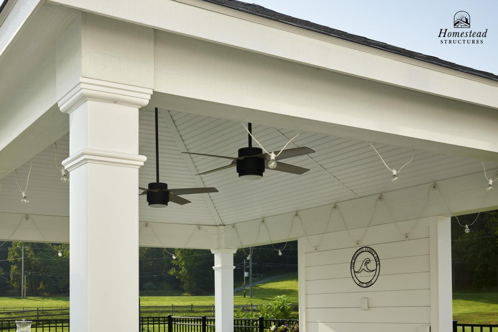 Covered outdoor patio with white columns, ceiling fans, string lights, and a sign with a wave logo indicating 'Homestead Structures'.