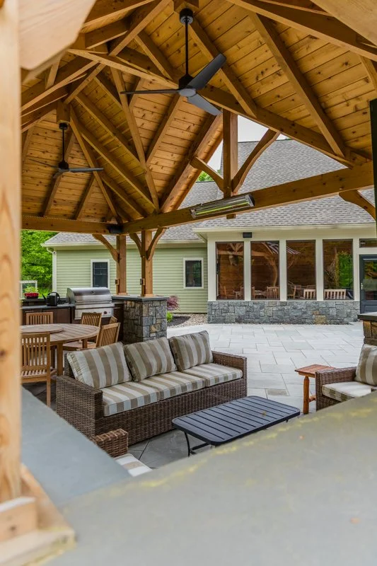 Outdoor covered patio area with a wooden ceiling, ceiling fans, and seating including a striped sofa and coffee table, with a house and garden in the background.