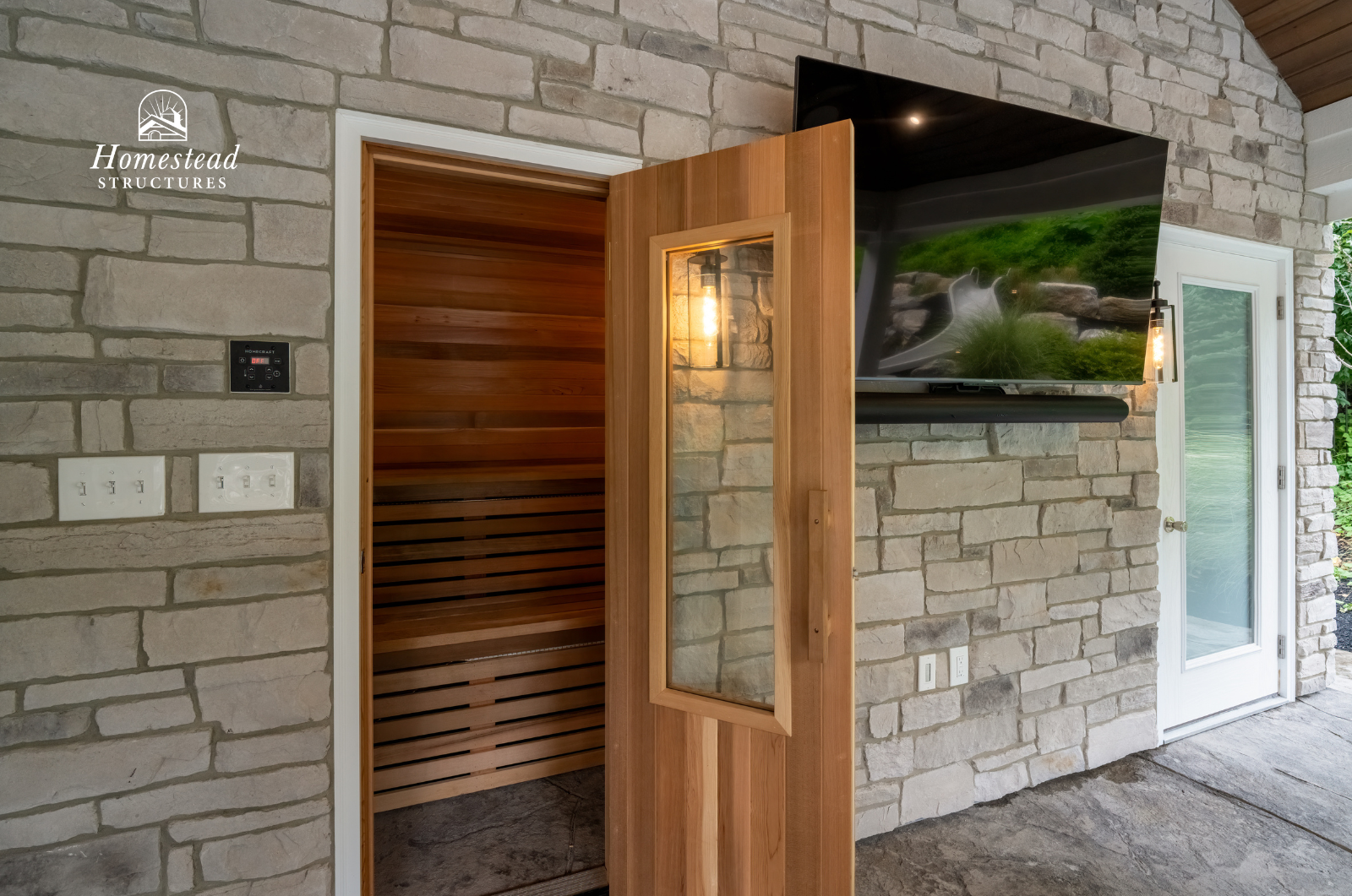 View of a wooden sauna room through an open door on a stone wall with a mounted TV, light fixtures, and a glass door leading outside.