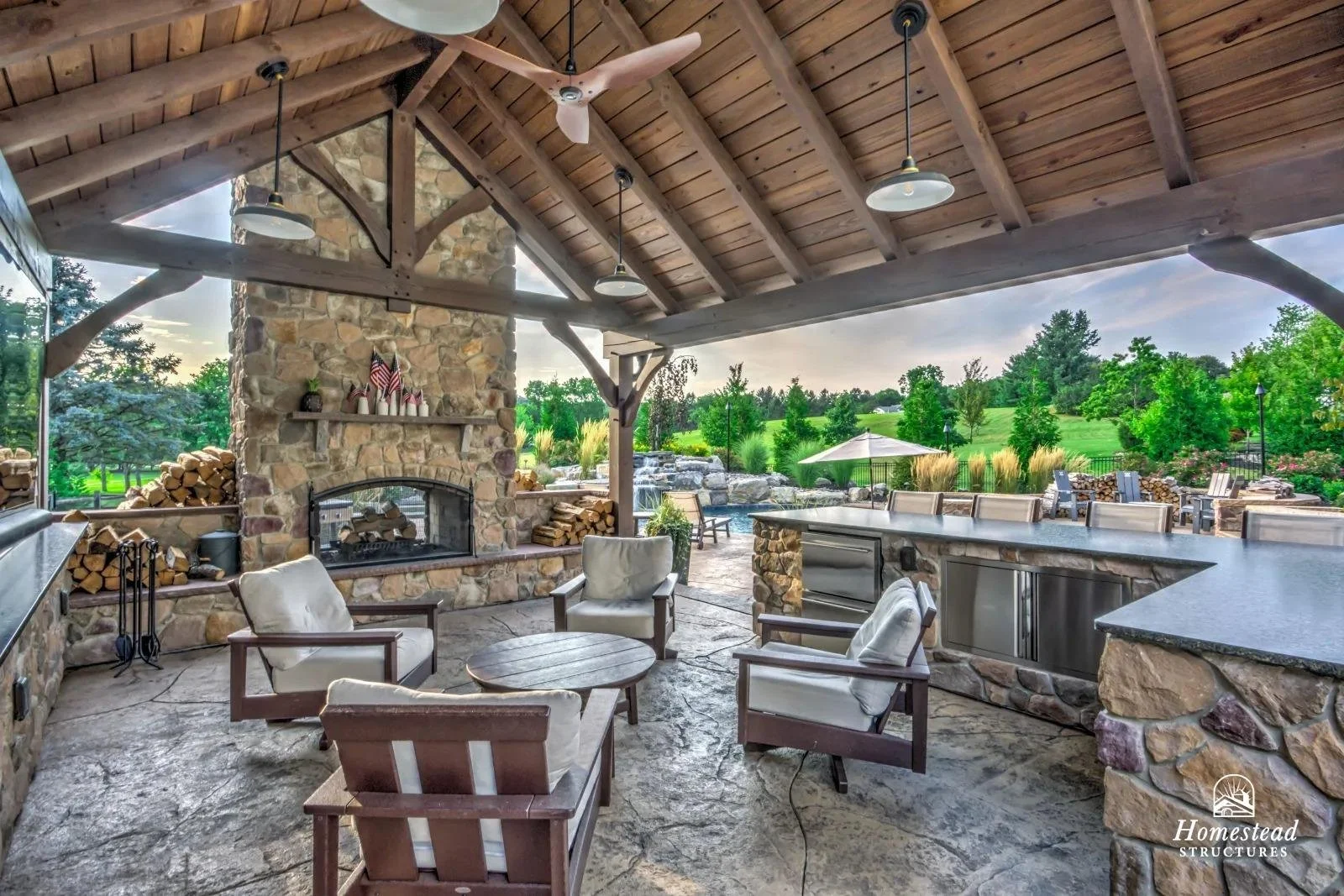 Outdoor covered patio with stone fireplace, ceiling fan, and hanging lights. Furniture includes cushioned chairs and a round table. The patio overlooks a lush green landscape with trees, a small waterfall, and a grill area.