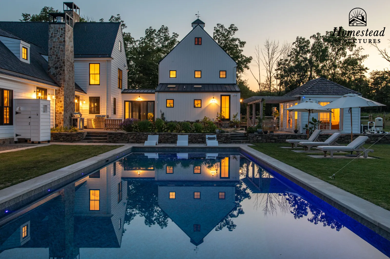 Luxury modern farmhouse with lit interior, outdoor swimming pool reflecting buildings and trees at sunset, with poolside lounge chairs and umbrellas