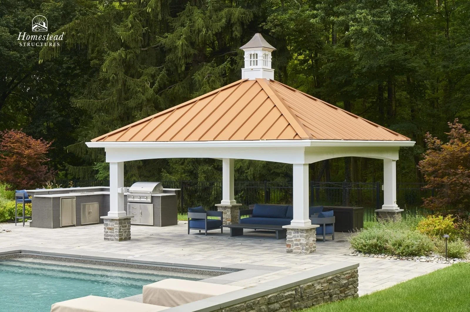 A backyard with a swimming pool and a covered patio area. The patio has seating, a built-in grill, and is surrounded by lush green trees.