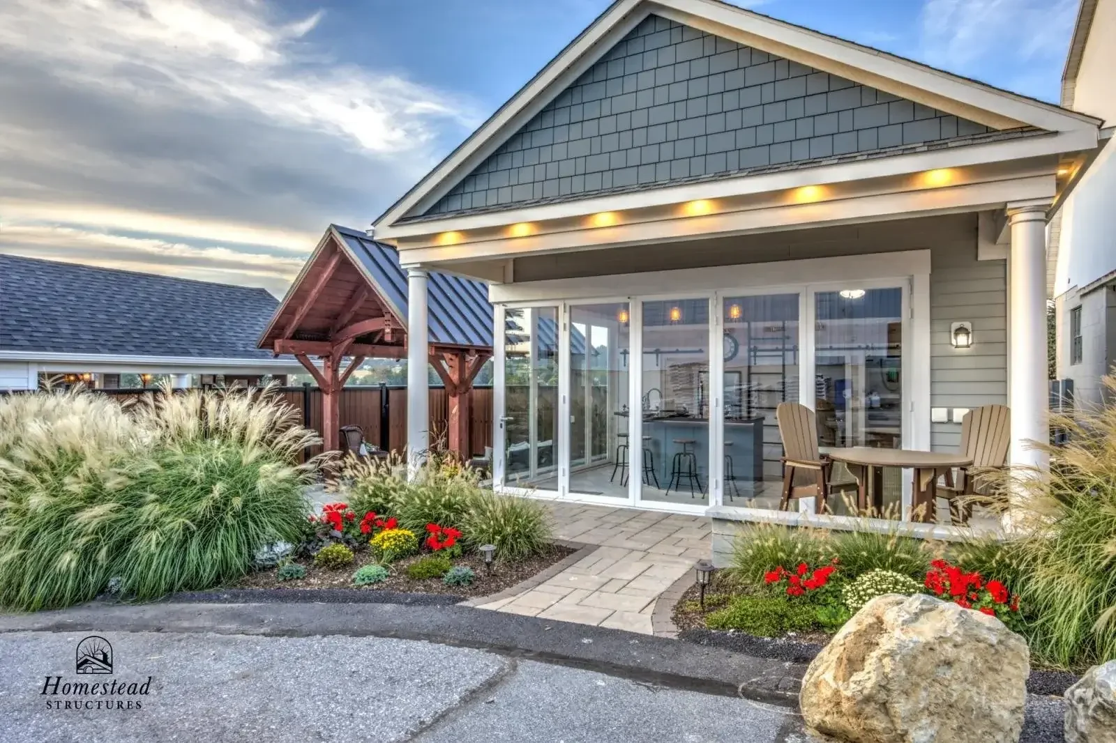 Backyard patio with garden, patio table, and chairs, surrounded by plants and flowers, with a house featuring a screened-in porch and small wooden gazebo.