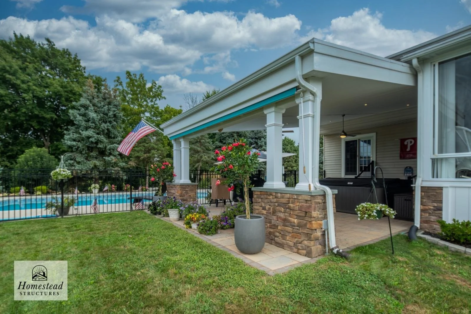 Backyard with a swimming pool, patio area with potted plants, flowers, and a covered seating area, surrounded by a black fence, with trees and a blue sky with clouds in the background.