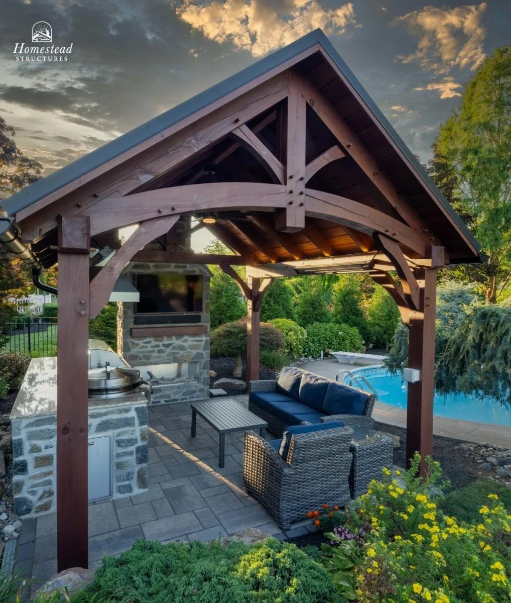 A backyard patio with a wooden gazebo, outdoor seating including a wicker couch with blue cushions, a matching chair, and a black table. There is a stone outdoor fireplace with a TV mounted on it, and a swimming pool in the background. The area is su