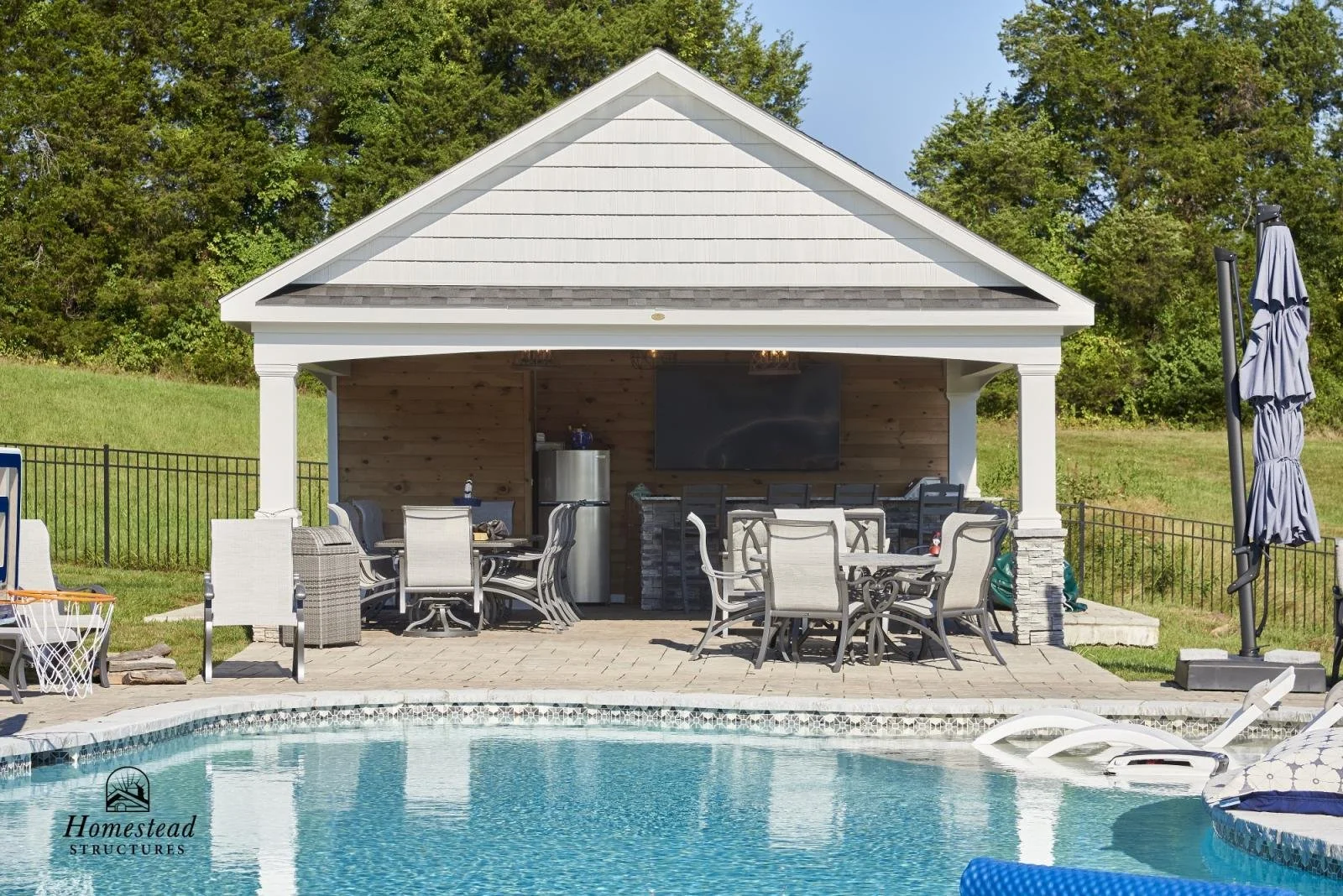 Outdoor pool area with a covered patio, seating, and a large flat-screen TV, surrounded by a grassy yard and trees.