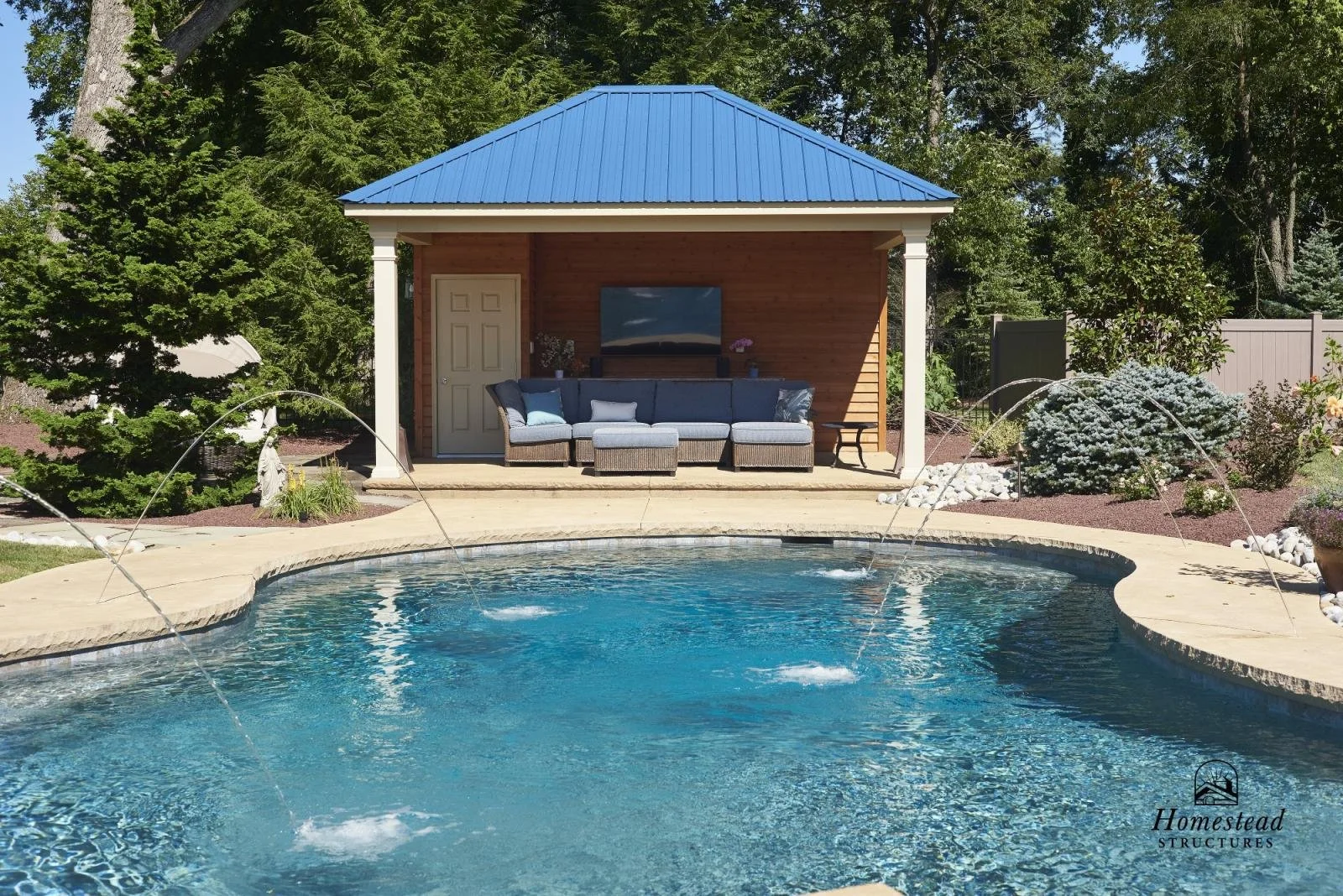 A backyard scene featuring a swimming pool with water features, a patio area with outdoor furniture, and a small wooden structure with a blue metal roof, surrounded by trees and landscaped bushes.