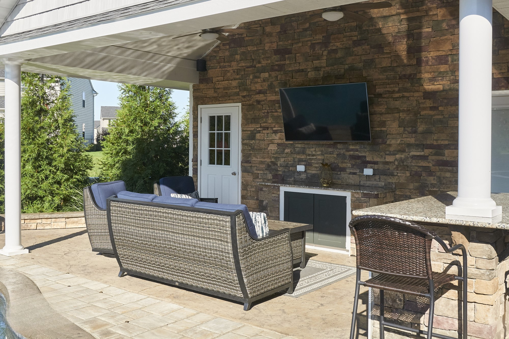 An outdoor patio area with wicker and cushioned furniture, a wall-mounted TV, a stone and brick wall, a white door, and a large umbrella providing shade.