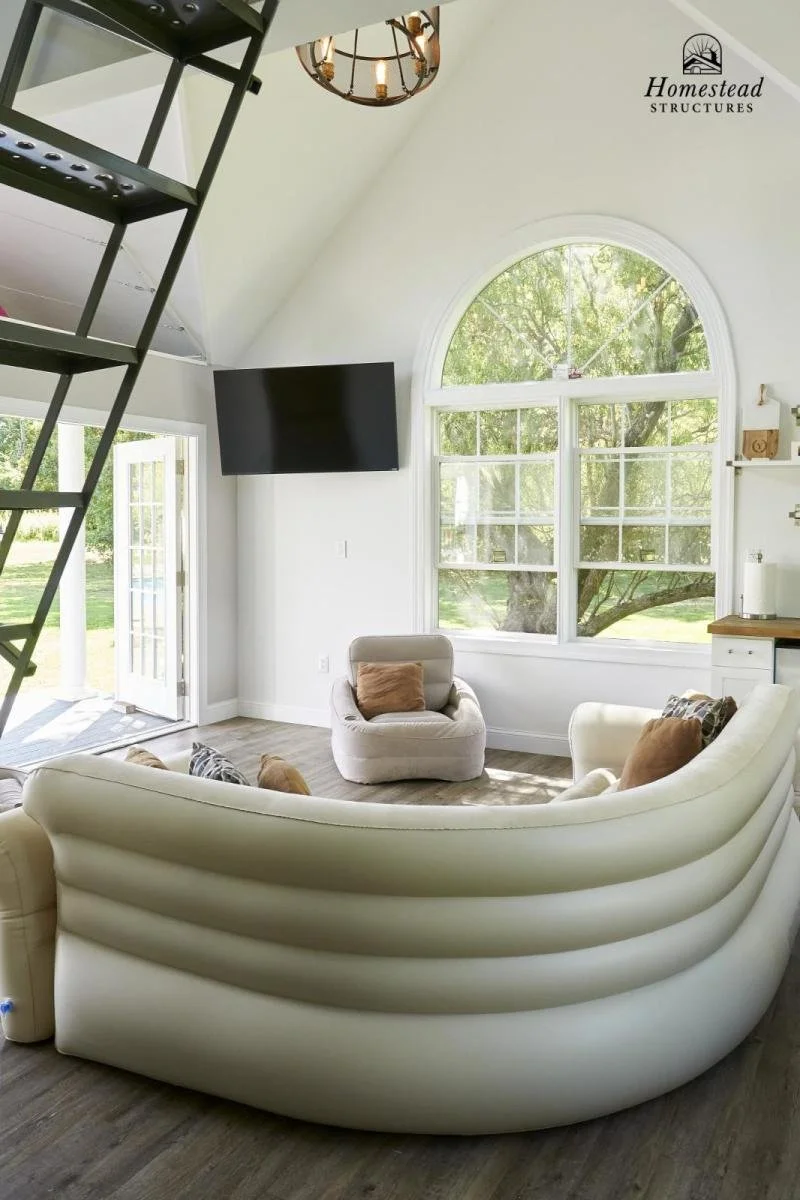 Modern living room with large arched window, white curved sofa, beige armchair, and wall-mounted flat-screen TV. There's a door leading outside, a hanging chandelier, and a view of greenery outside.