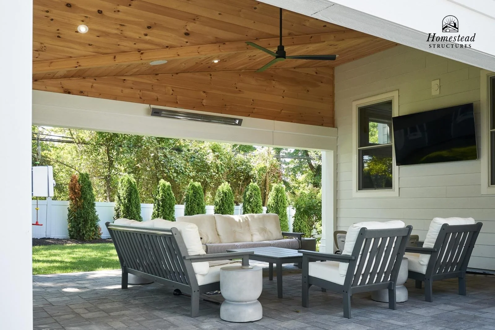 Outdoor patio with cushioned seating, coffee table, and white stone side table under a wooden ceiling with ceiling fan, surrounded by greenery and a white fence