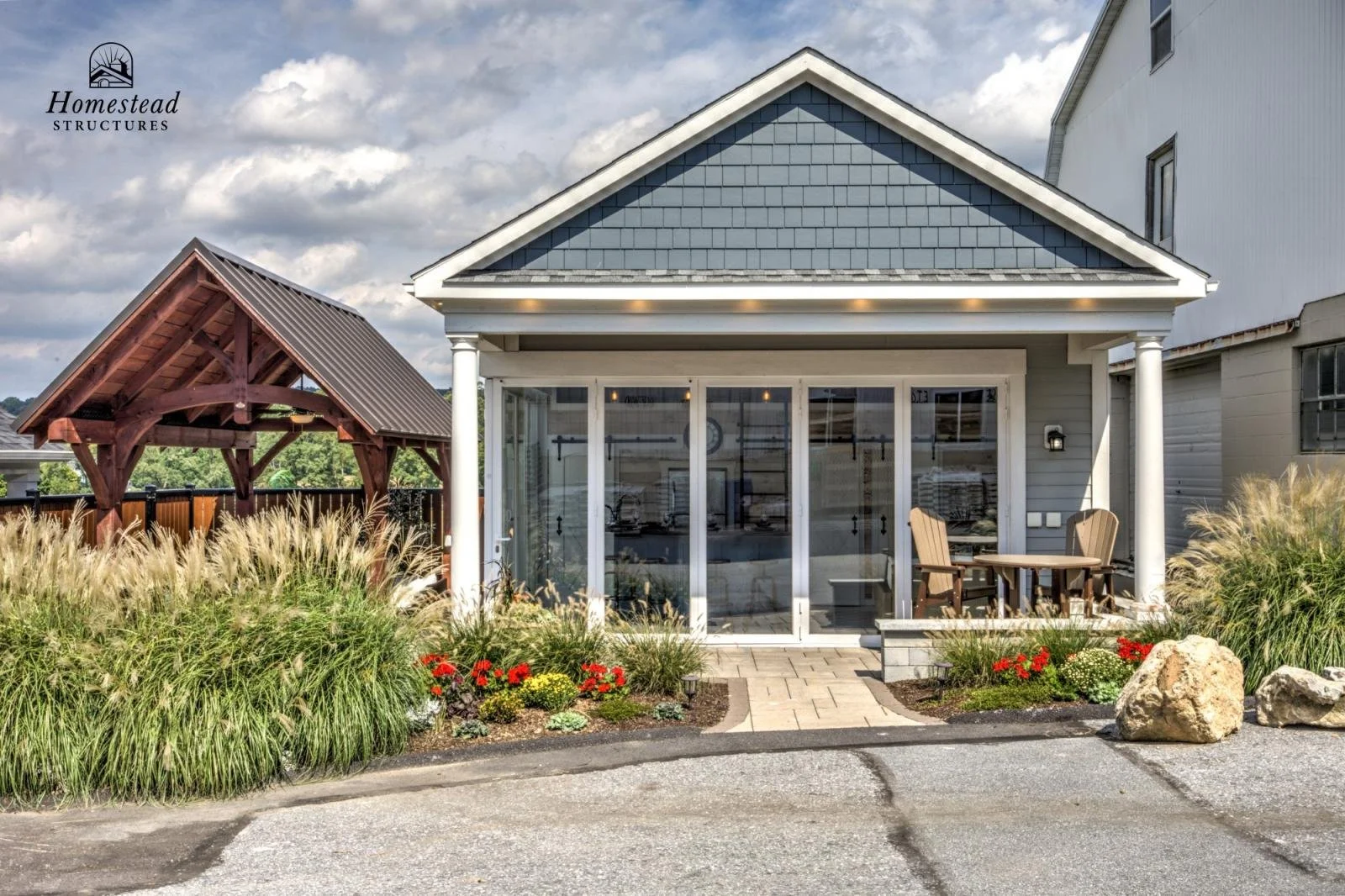 Front view of a modern house with a glass porch and outdoor seating area, surrounded by landscaped garden plants and rocks, under a partly cloudy sky.