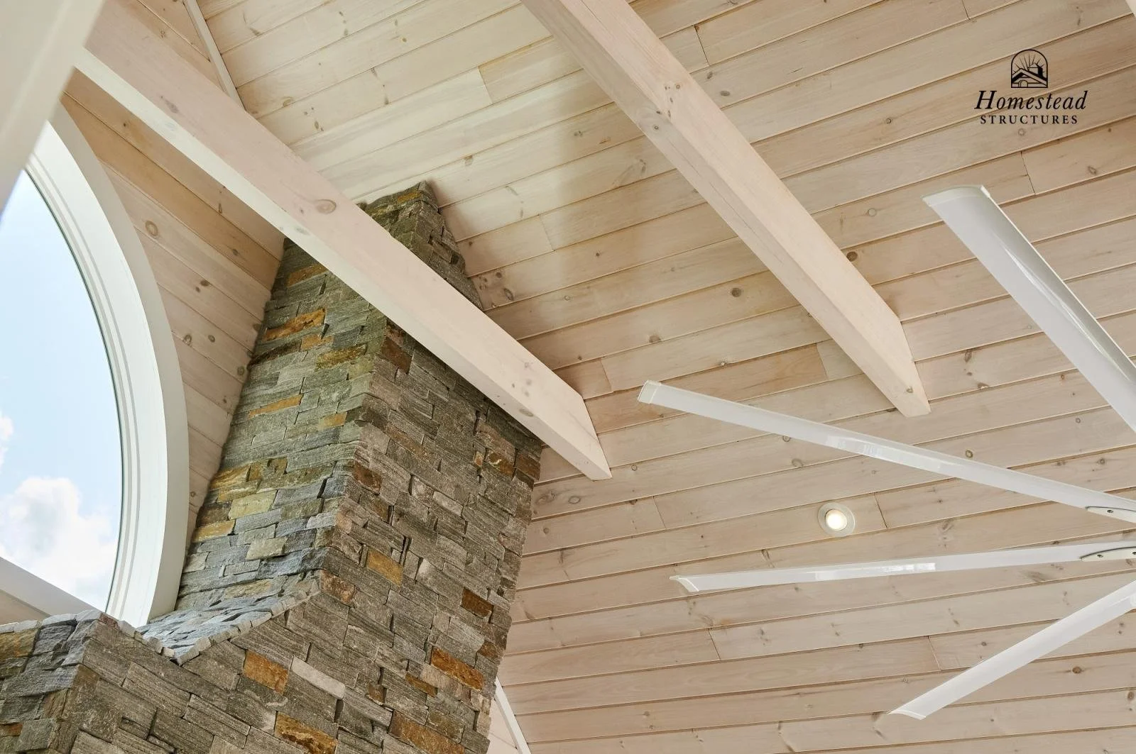 Interior ceiling with light wood paneling, a stone chimney, and modern white hanging light fixtures, with a large arched window showing a blue sky with clouds.
