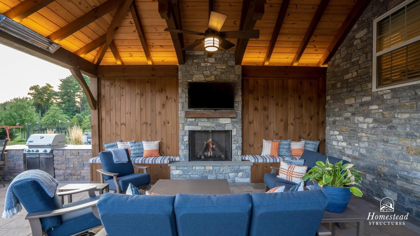 Outdoor covered patio with blue cushioned seating, a stone fireplace, a mounted TV, wooden ceiling with a ceiling fan, and a brick and wood wall. There is a potted plant on a side table and a grill on the left outside the patio.