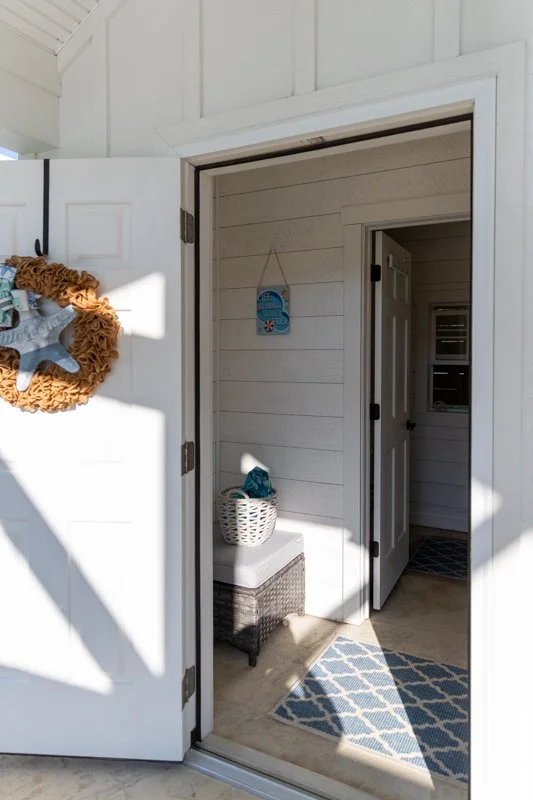 Open door to a room with white walls and a wicker basket with blue towel inside, a small bench, a blue patterned rug, and a decorative blue wall hanging.