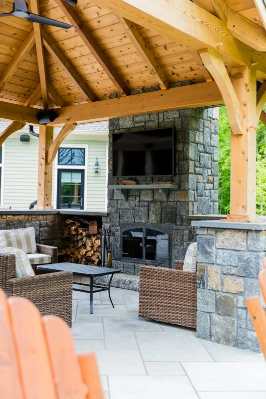Outdoor patio with a stone fireplace, a TV mounted above, wooden beams supporting a roof, and wicker furniture.