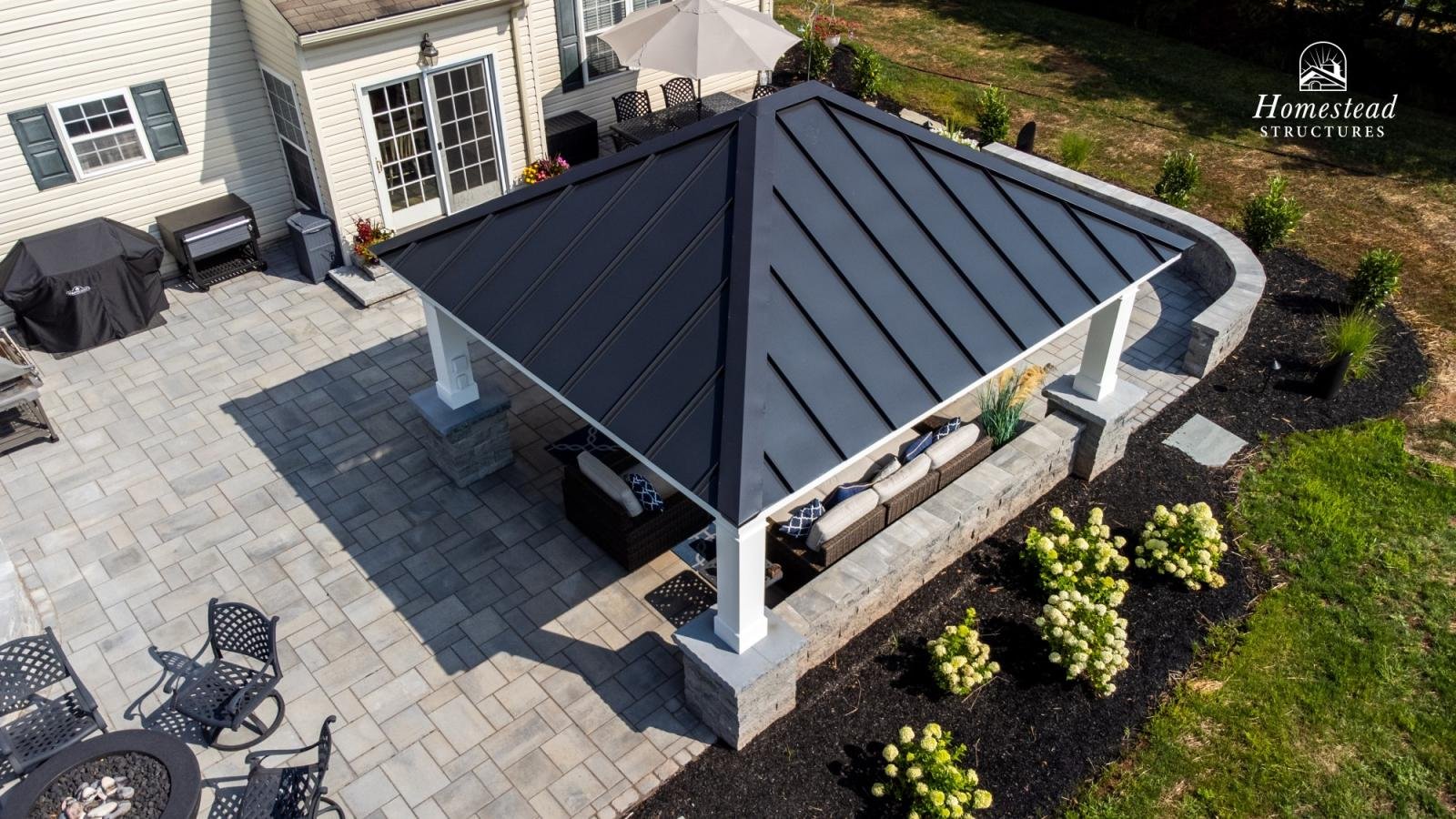 A backyard patio with a metal roof gazebo, outdoor furniture, a barbecue grill with cover, potted plants, and landscaped garden with white hydrangeas.