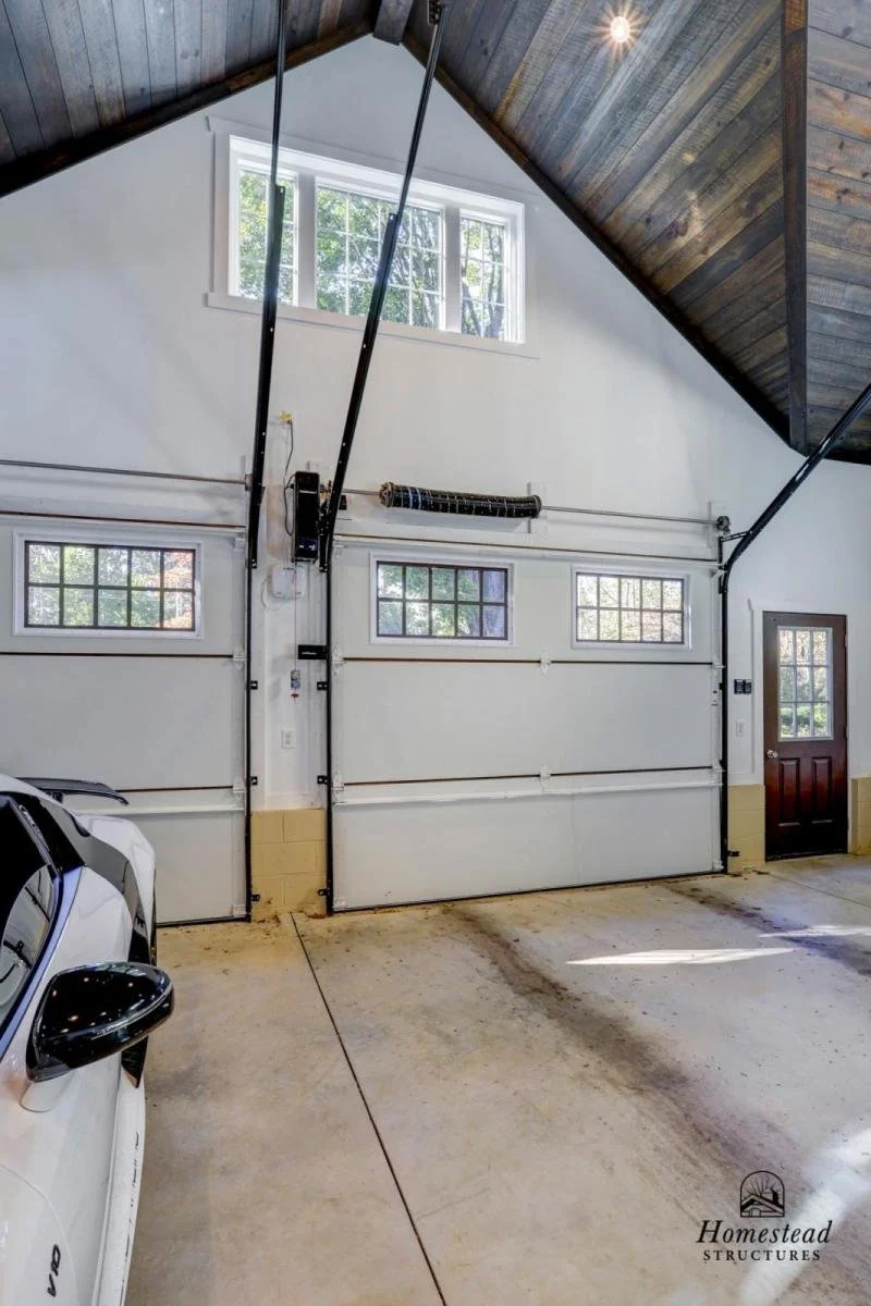 A garage with a white sectional door featuring three small windows and a side door, a high ceiling with wooden paneling, and an overhead garage door opener system. A white vehicle's side mirror is visible in the lower left corner, and there's a logo 