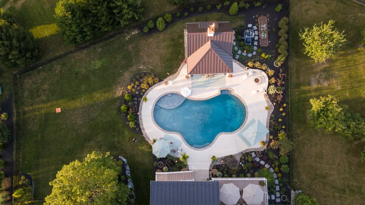Aerial view of a backyard with a kidney-shaped swimming pool, surrounding patio, umbrellas, and landscaped garden with trees and bushes.