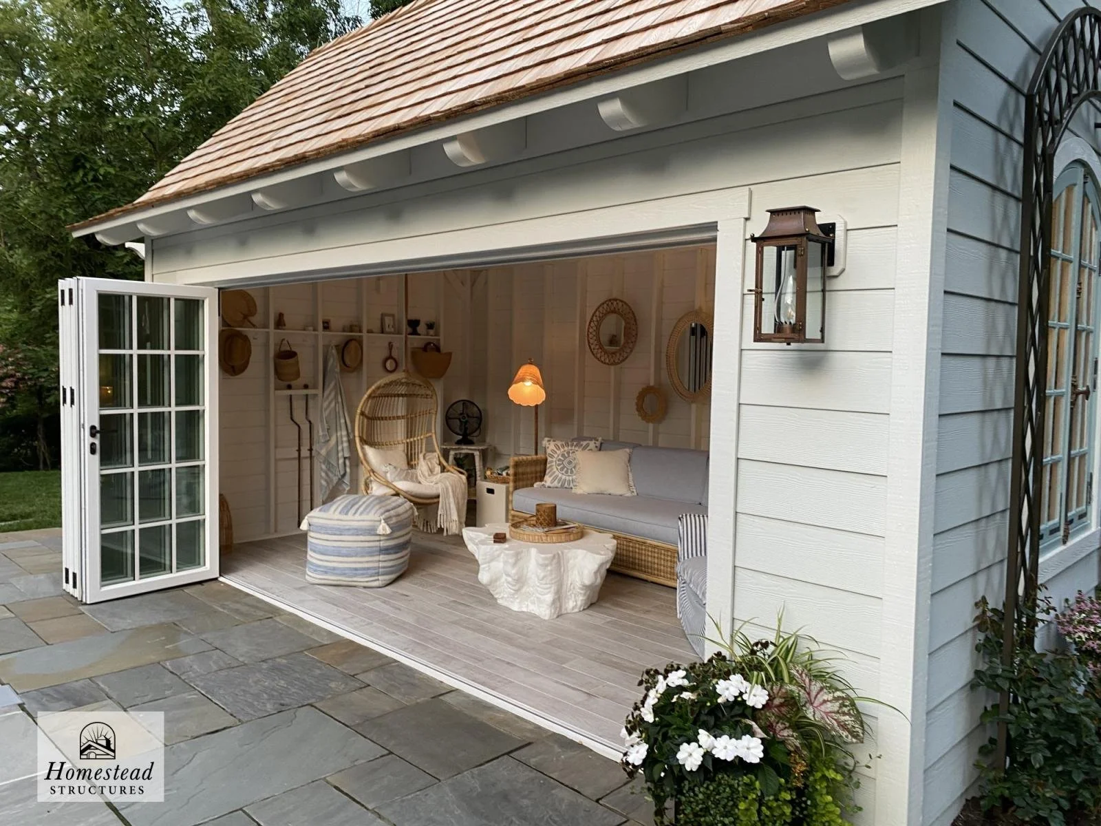 Decorative outdoor living space inside a white wooden garden shed with open bi-fold doors, featuring wicker chairs, a gray sofa, a round coffee table, and outdoor decor such as mirrors, curtains, and a hanging lamp.