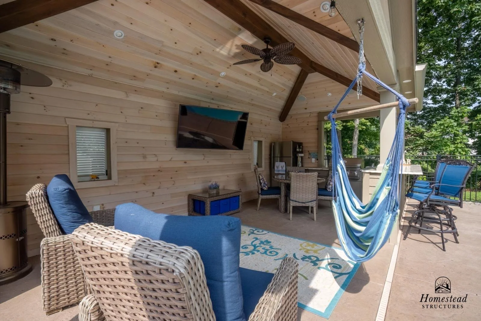 Covered outdoor patio with wicker and fabric seating, a hanging hammock chair, a flat-screen TV, a ceiling fan, and a bar area, surrounded by trees.