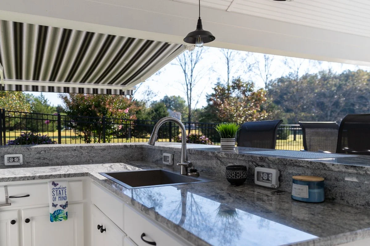 Outdoor kitchen with granite counter, black chairs, potted plant, and view of trees and fence outside.