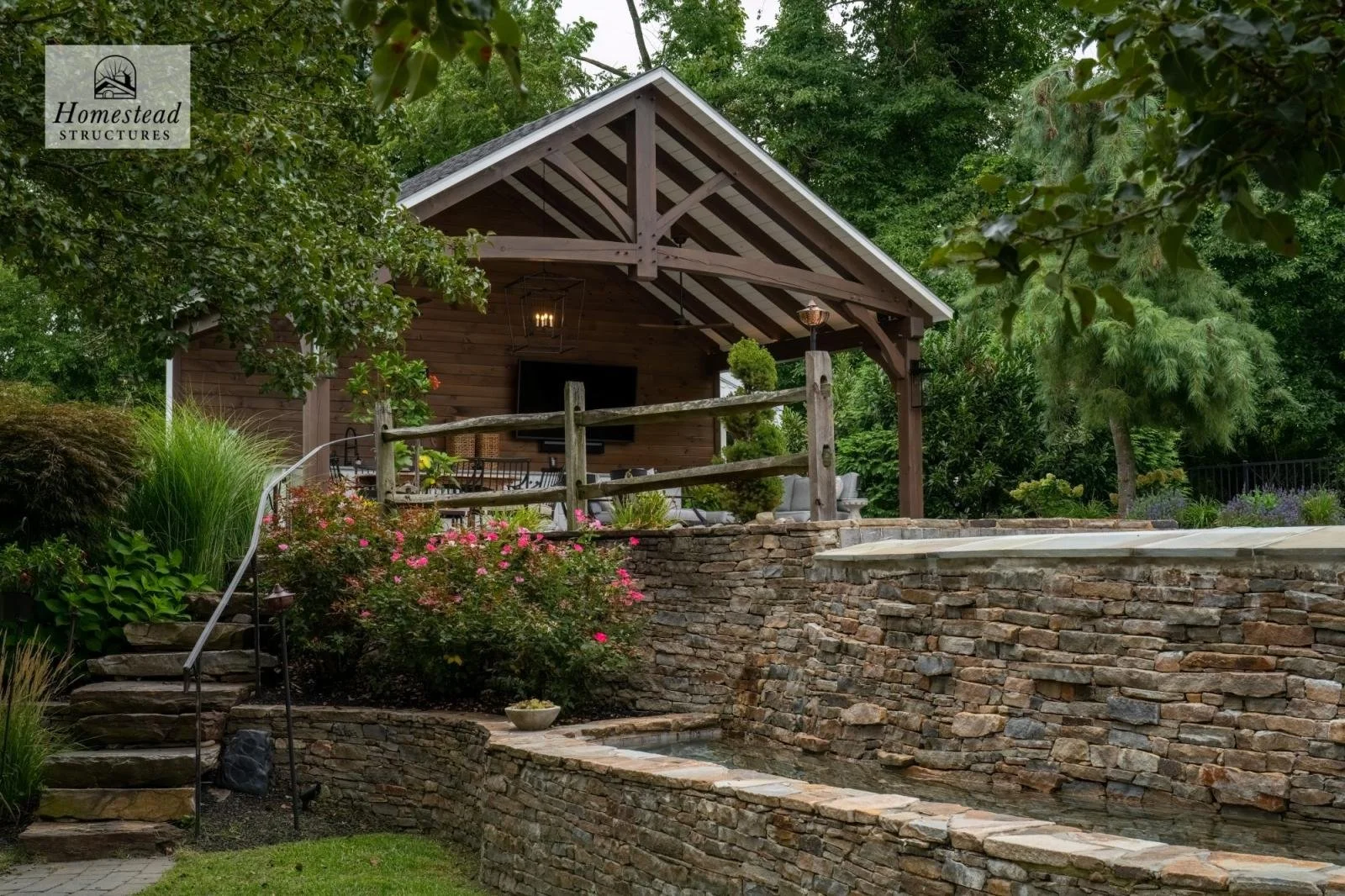 A backyard with a stone retaining wall, a wooden pergola with outdoor seating, lush greenery, flowering bushes, and trees.