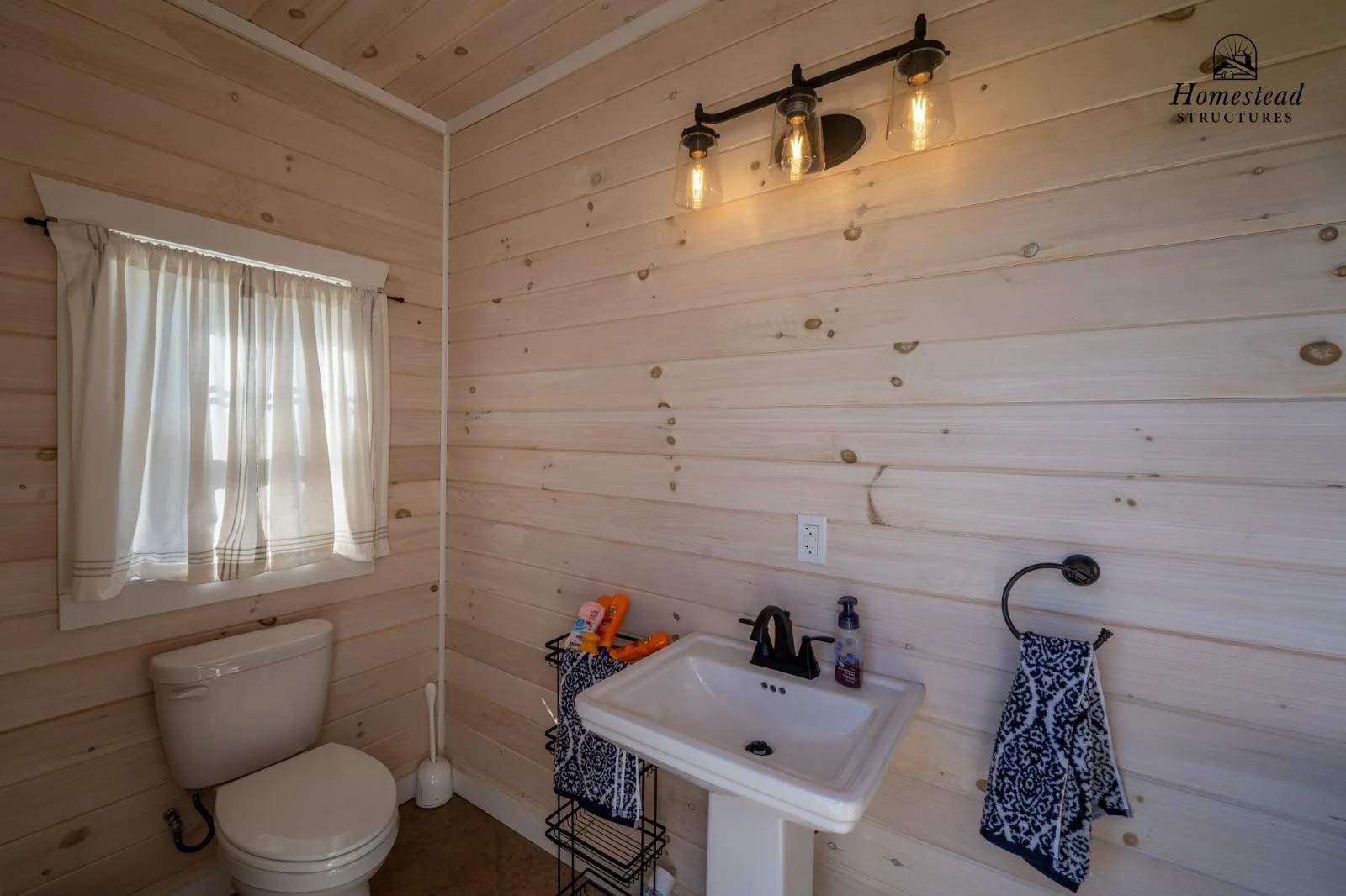 Small bathroom with wood-paneled walls, a window with white curtains, a toilet, a small sink with a black faucet, a black towel ring with a blue patterned towel, a black wire shelf with toiletries, and a ceiling light fixture with three warming bulbs