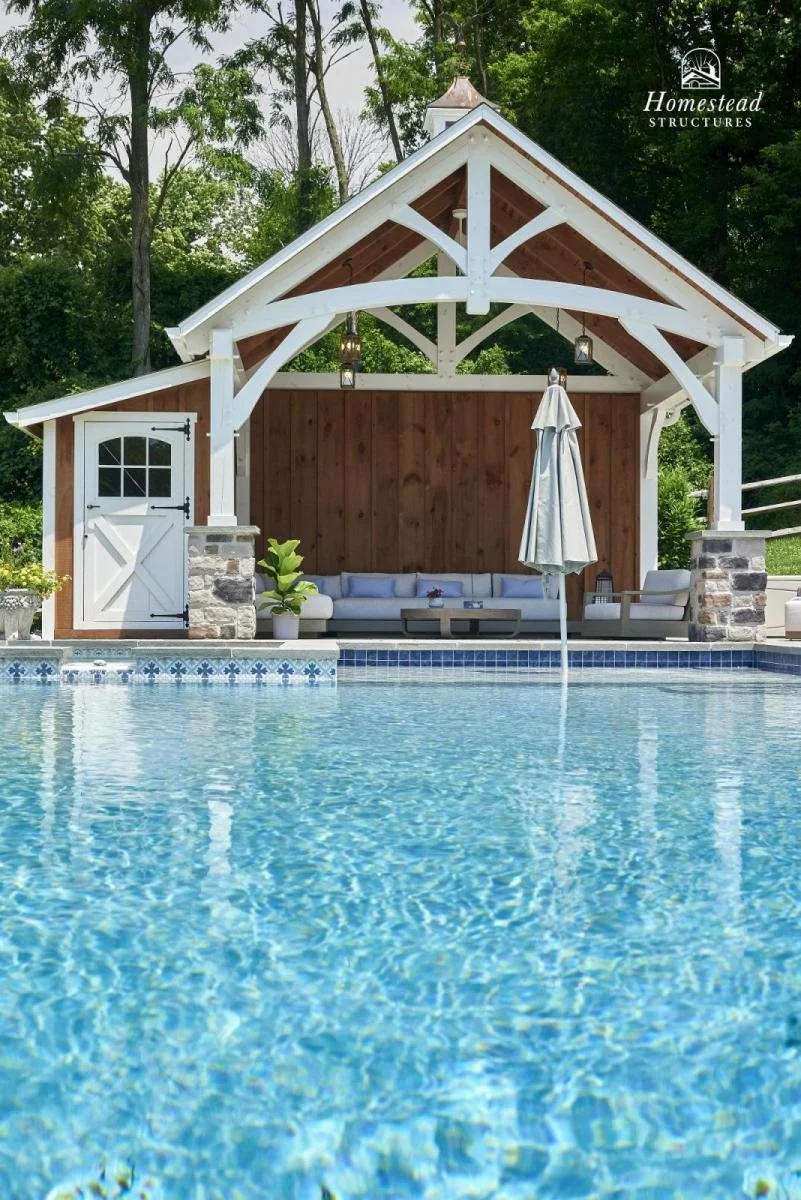 A backyard scene with a swimming pool and a poolside cabana with white and wood accents, hanging lanterns, a cushioned sofa, a closed umbrella, and lush green trees in the background.