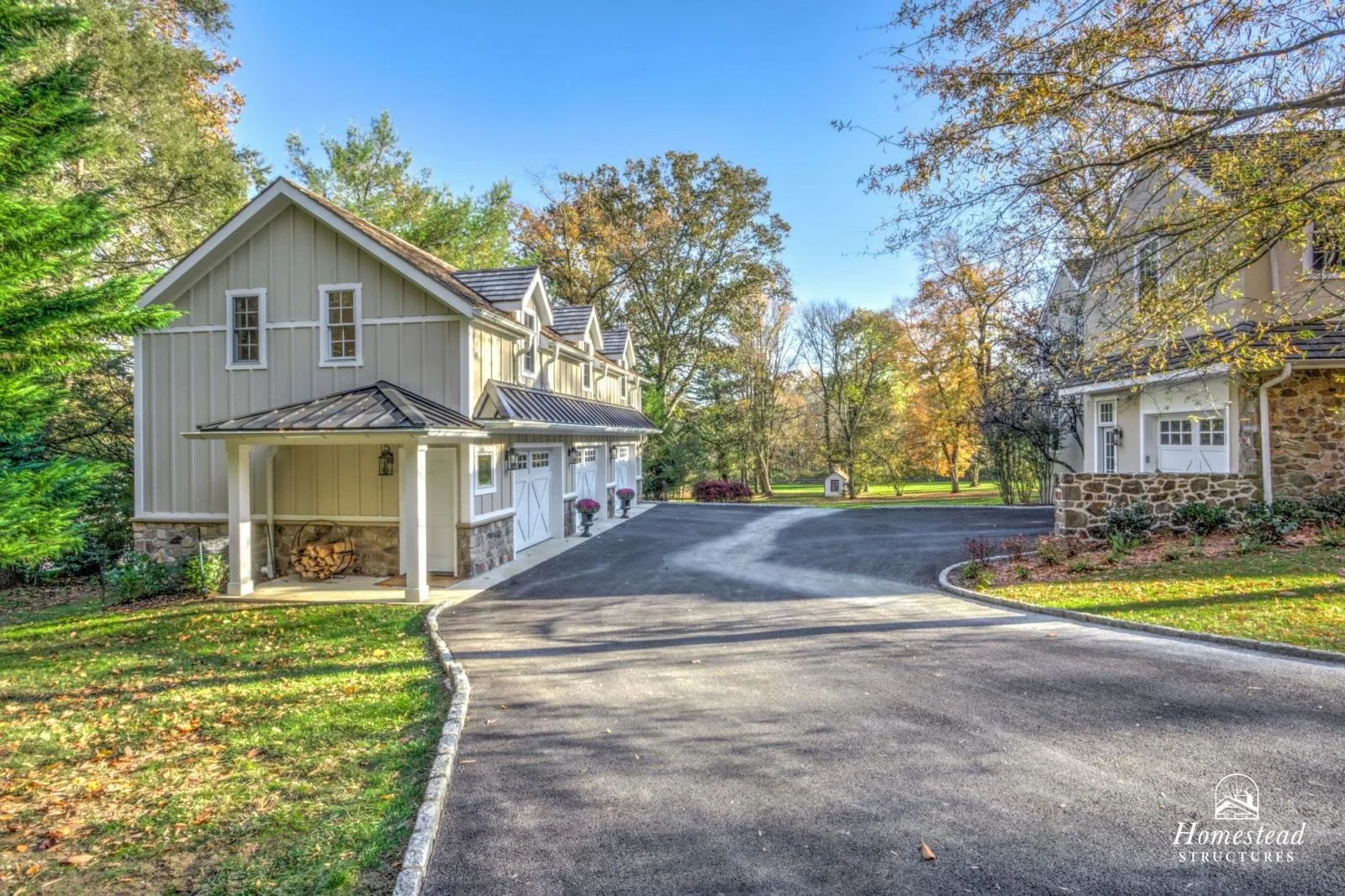 A gravel driveway curves past two houses with white and stone exteriors, surrounded by trees with autumn foliage, under a clear blue sky.