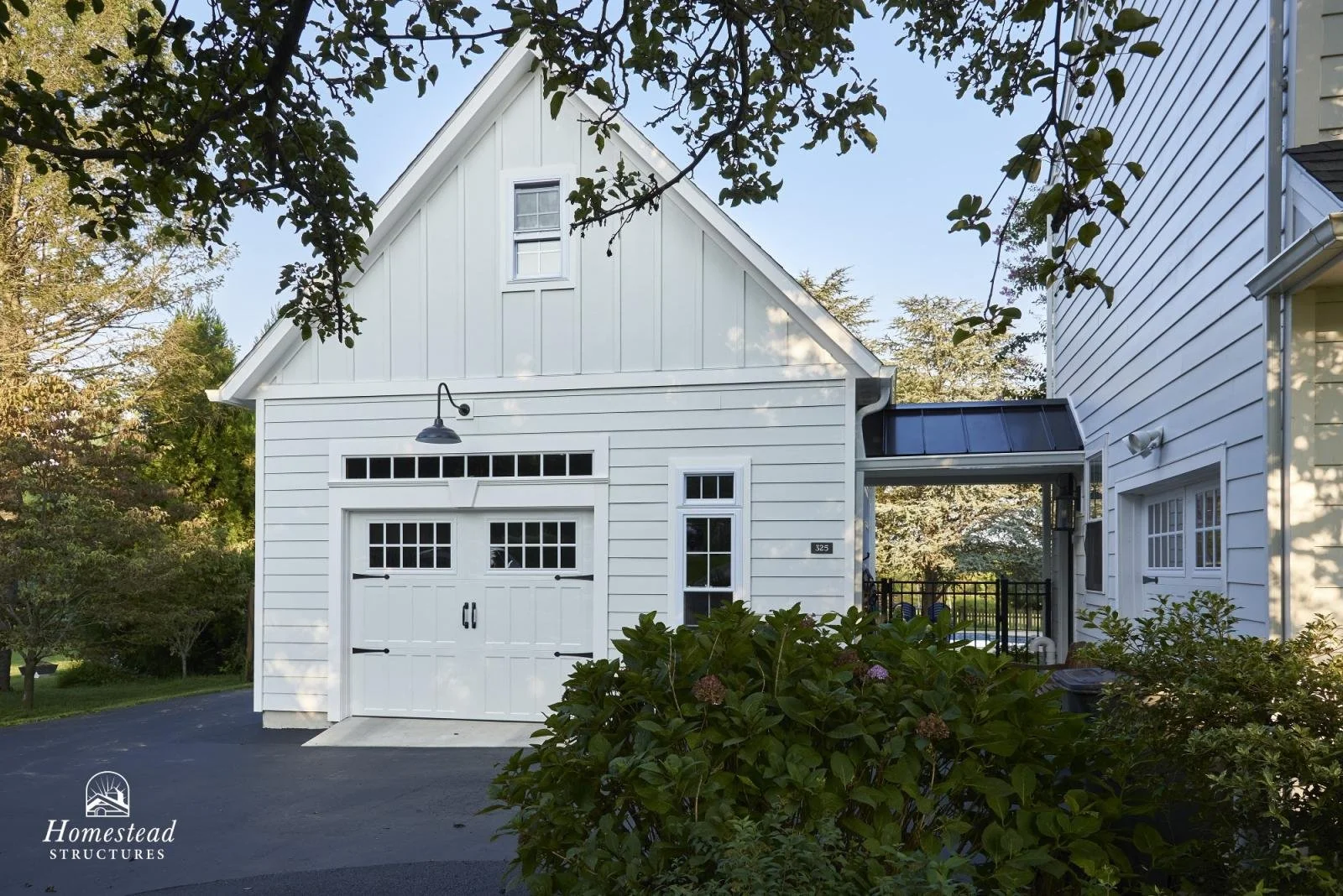 A white barn-style garage with black hardware, installed on a paved driveway, with a small window and porch light. Adjacent to a white house with horizontal siding, surrounded by trees and bushes, under a clear blue sky.