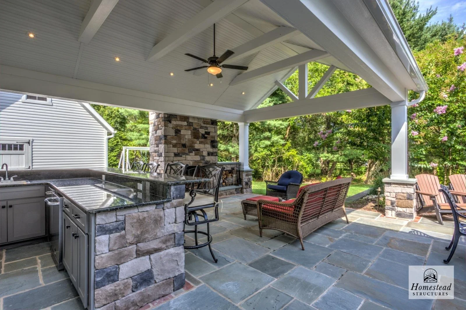 Covered outdoor patio with stone flooring, seating area, built-in outdoor kitchen with granite countertops, ceiling fan, and surrounding trees with pink flowers.