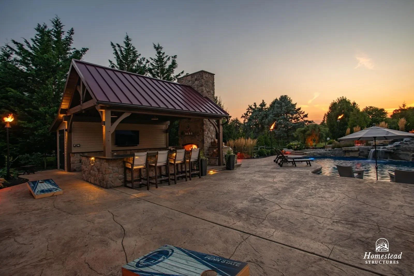 A backyard patio during sunset with a stone and wood outdoor bar with barstools, pergola, fireplace, pool with lounge chairs, and tiki torches.