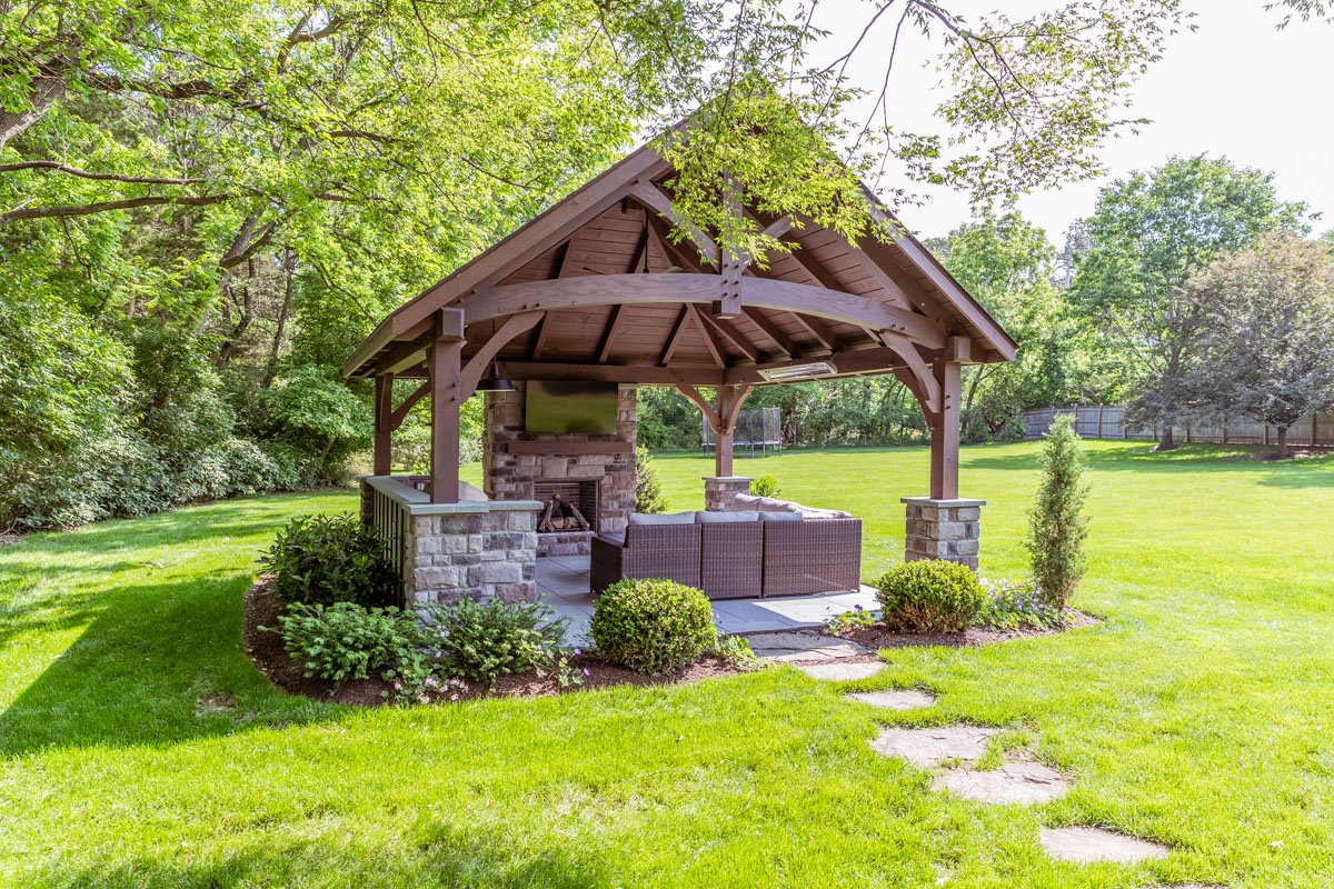 A gazebo with a wooden roof and stone columns in a lush backyard with green grass and trees, featuring outdoor furniture and a fireplace.