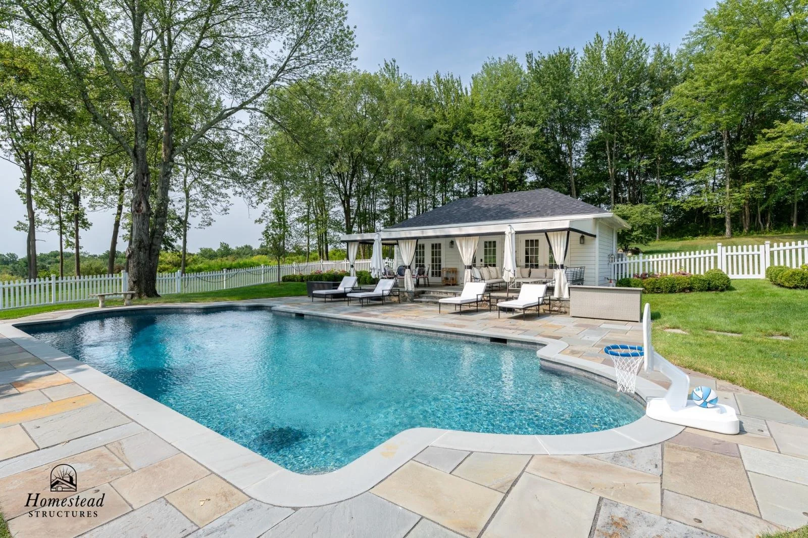 A backyard with a swimming pool, poolside lounge chairs, a seating area under a covered patio, a white fence, green grass, and trees in the background.