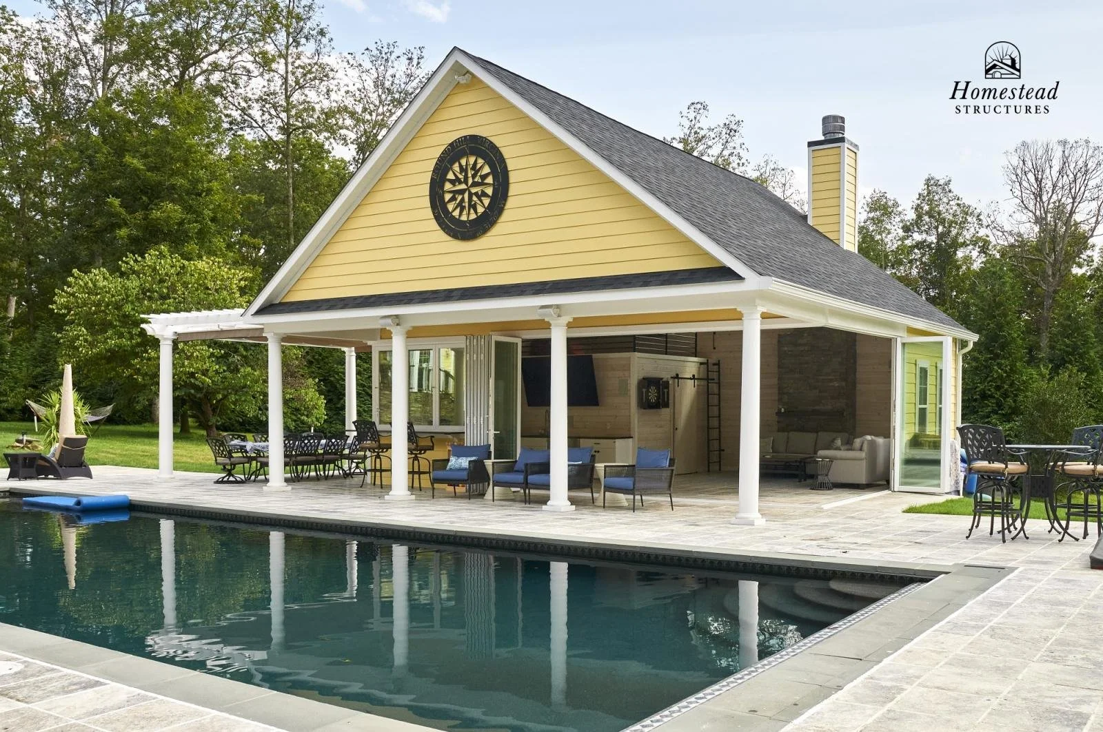 A yellow house with white columns, a gray roof, and a chimney, situated next to a swimming pool with outdoor furniture.