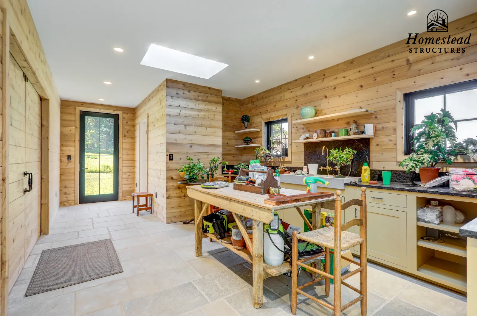 Interior of a rustic kitchen with wooden walls and a stone floor, featuring a small kitchen island, open shelves with decor, a window, and a door leading outside.