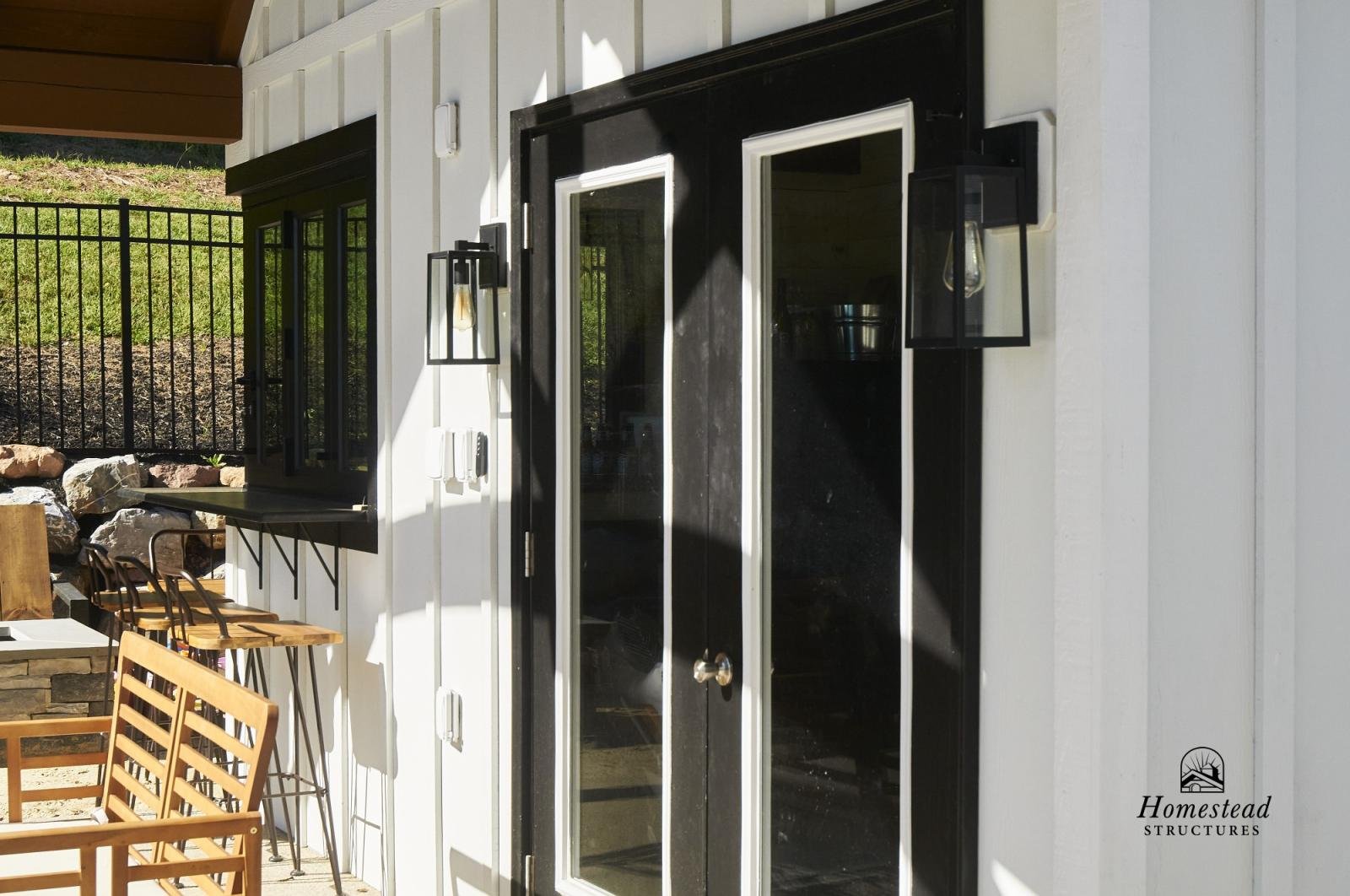 Exterior view of a building with black double glass doors, black outdoor wall-mounted lantern lights, a small window shelf with a black frame, and a white wall with visible siding. A metal fence and green lawn are in the background, and part of a woo