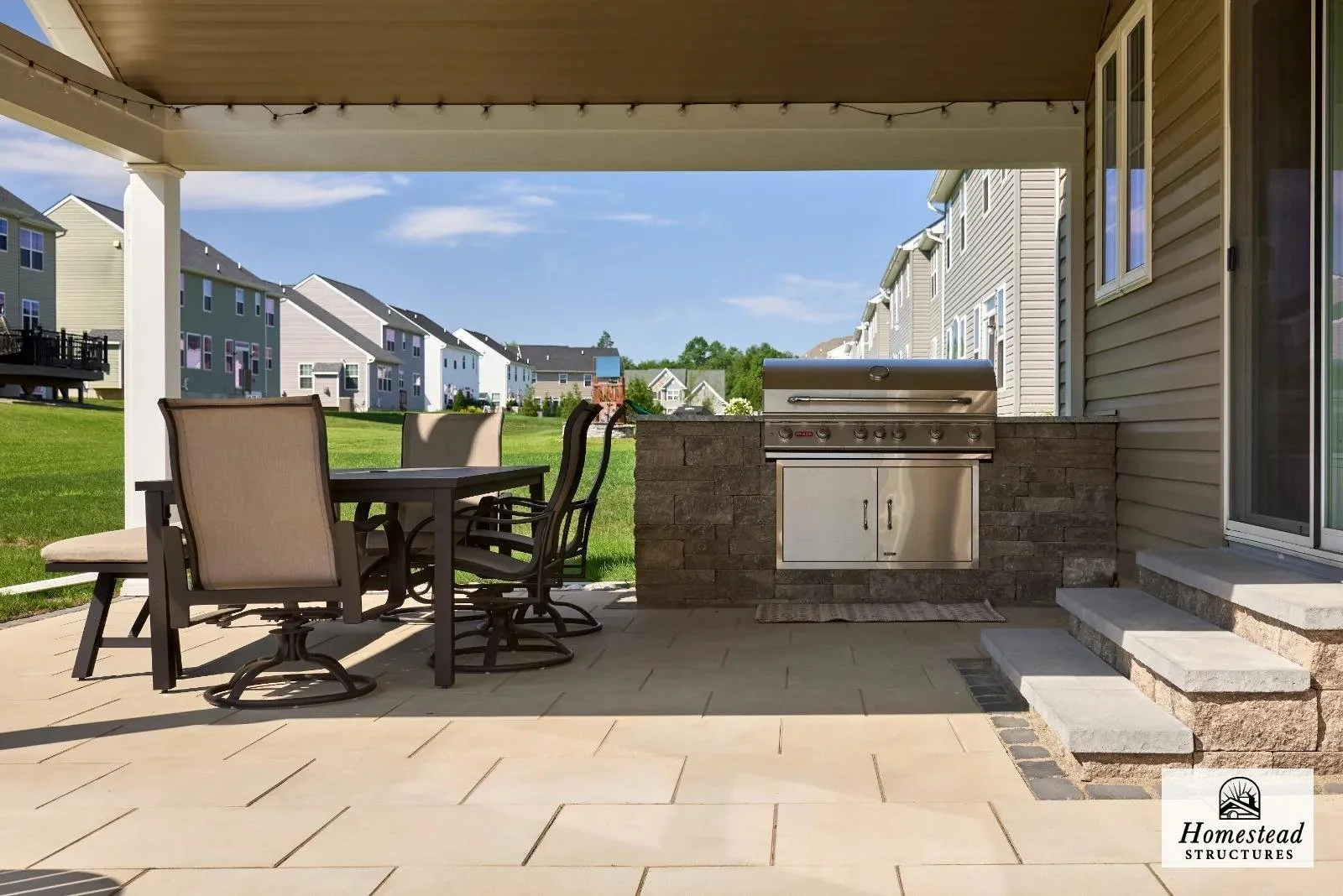 Backyard patio with a dining table, chairs, and a built-in stainless steel grill, with a grassy area and modern suburban houses in the background.