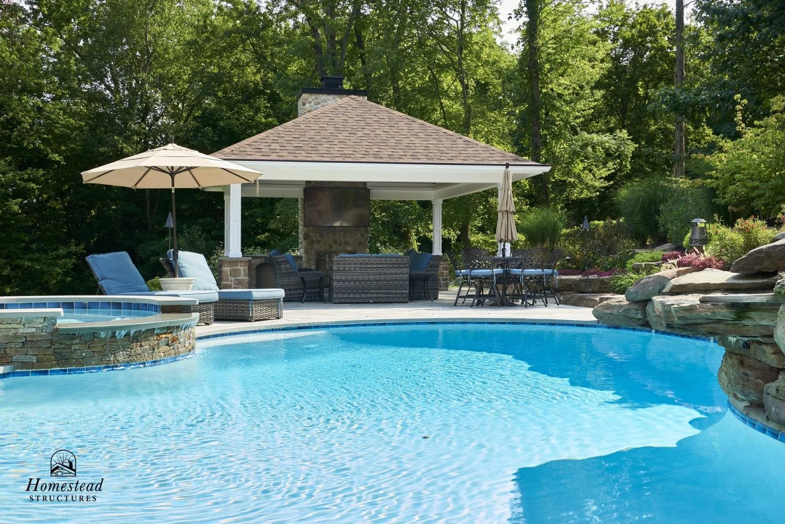 A backyard swimming pool area with lounge chairs, umbrellas, a pergola with seating and a television, surrounded by trees and landscaping.
