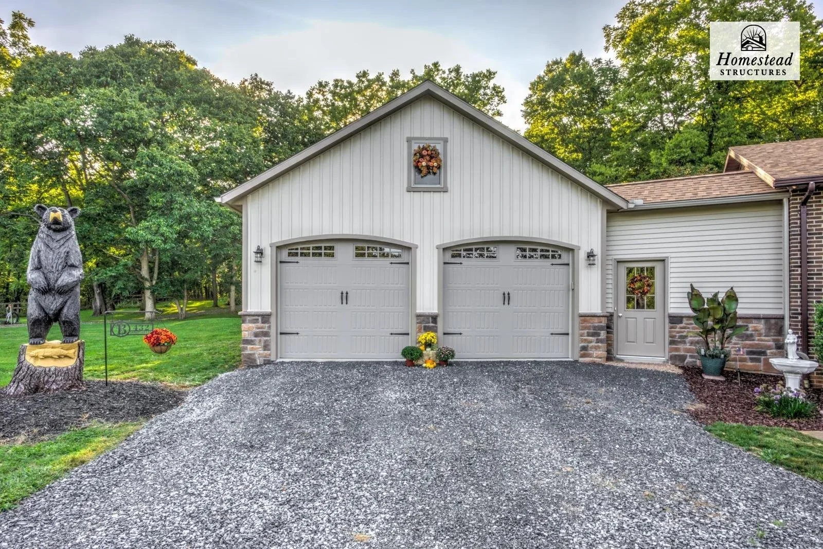 Front view of a white garage with two doors, stone accents, and decorative pumpkins in front. To the left, a bear statue with a yellow saddle stands on a tree stump. To the right, potted plants, a small door with a wreath, and a birdbath. Lush green 