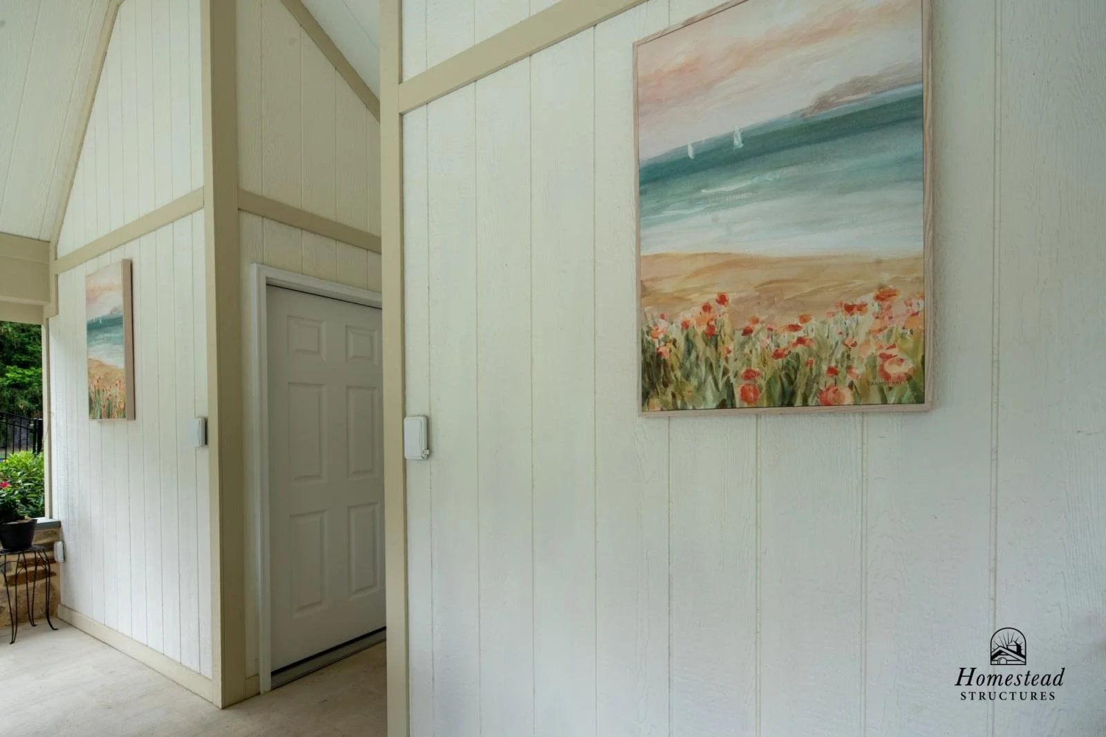 Interior view of a room with white paneled walls, a door, and landscape paintings hanging on the wall, featuring flowers, a beach, and sailboats.