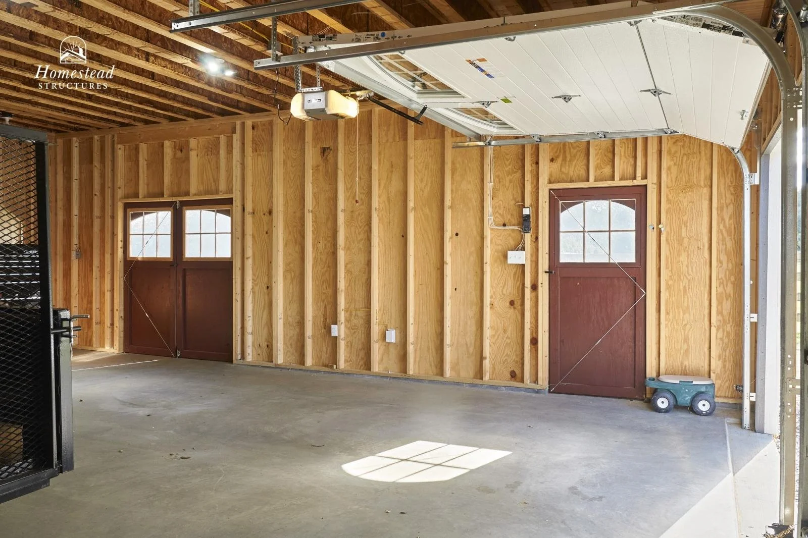 Interior of a newly constructed garage with unfinished wooden walls, a concrete floor, a partially open garage door, a small window, and a large with a pull-up door system. There is a small toy wagon and some garage door openers attached to the ceili