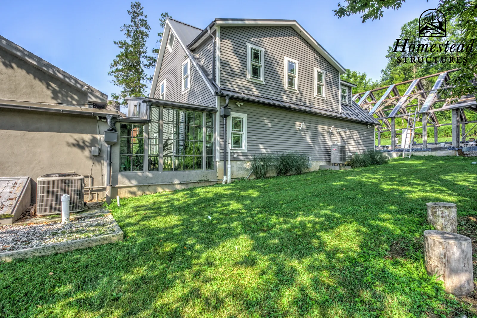 The image shows the backyard of a house with a well-maintained grassy lawn, several trees, and a partially constructed wooden structure on the right side. There is a small porch with a glass enclosure attached to the house, which has gray siding and 