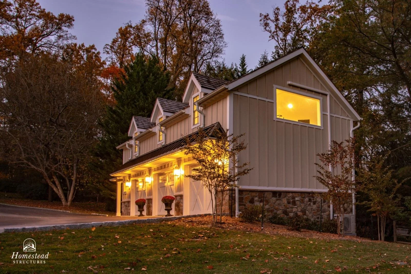 A modern three-story house with exterior lighting, multiple windows, on a slight hill with colorful autumn trees in the background, and a stone foundation.