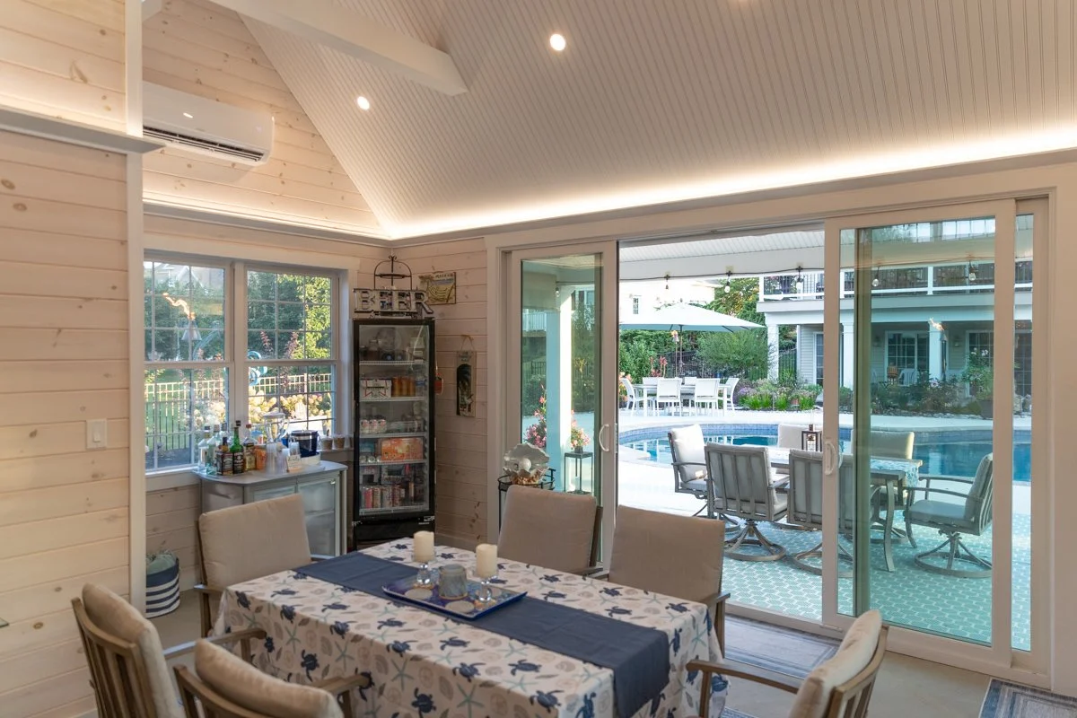 Indoor dining area with a wooden table covered with a patterned tablecloth, six chairs, and large sliding glass doors leading to outdoor patio with a pool and garden.